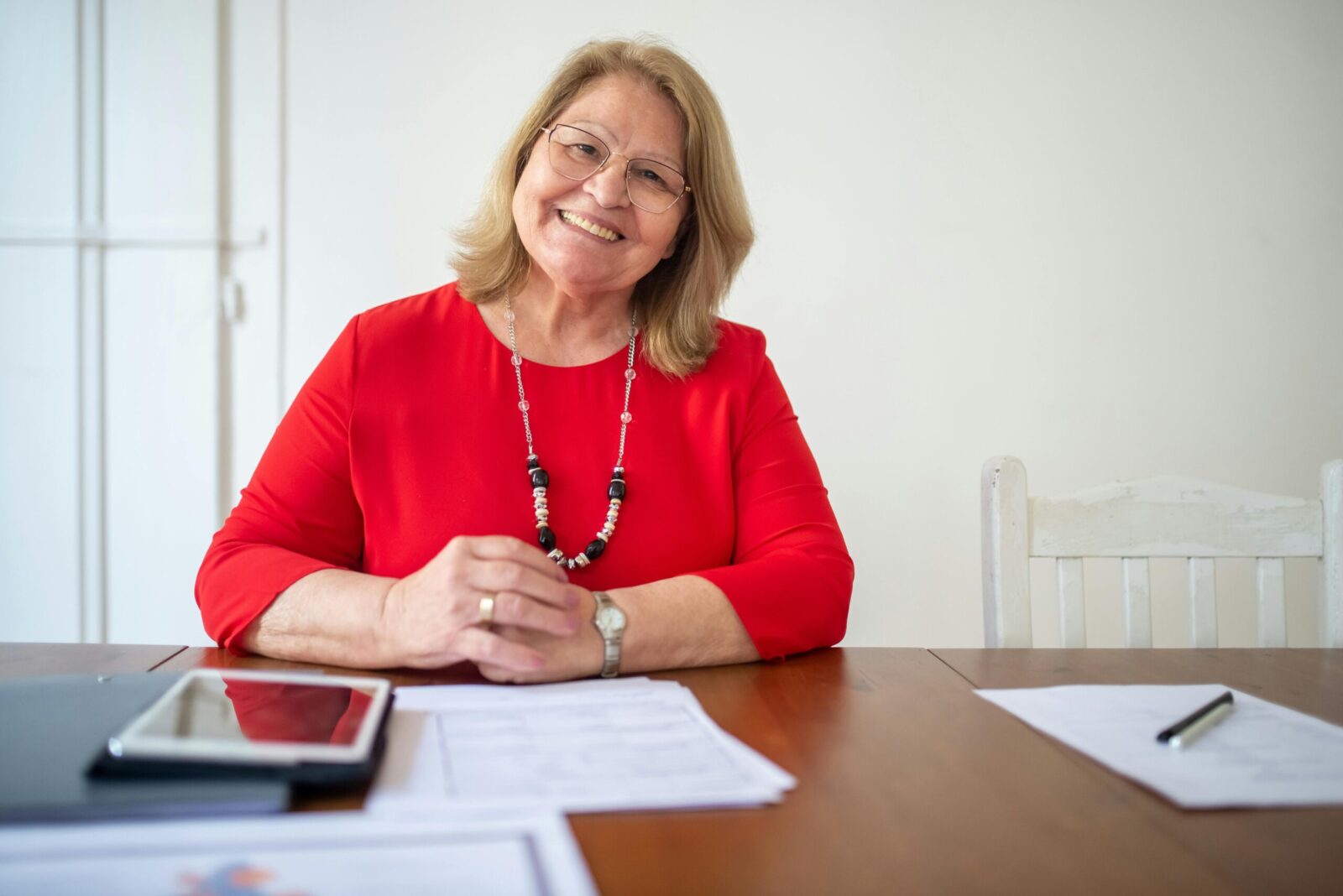Warm and approachable senior woman sitting at a desk with documents, wearing a bright red top.